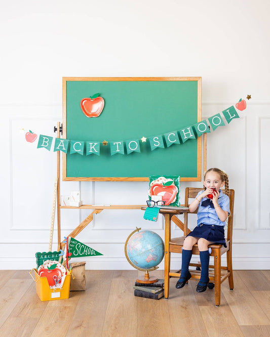 Young girl sitting at a desk with a 'Back to School' banner and green chalkboard in the background.
