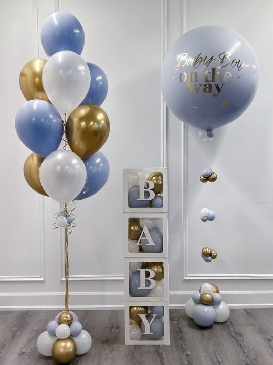 A baby shower decoration set featuring a cluster of light blue, white, and metallic gold balloons on the left, a large light blue balloon with gold foil text reading 'Baby Boy on the way' on the right, and a stack of three clear blocks displaying the letters 'B', 'A', 'B', 'Y' with gold and blue balloons inside.