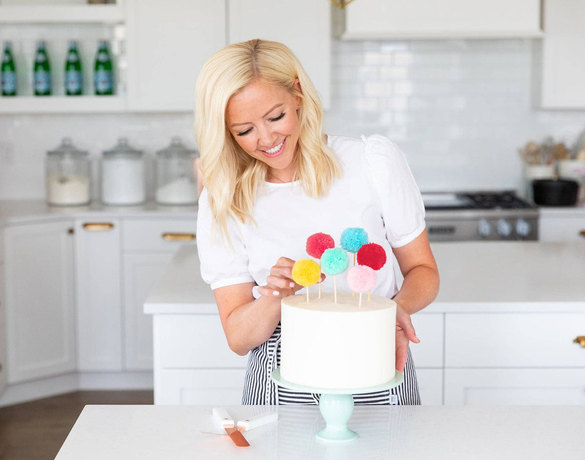 Colourful pom pom cake toppers decorating a white birthday cake on a mint green cake stand. Pick up in Toronto + Canada-wide shipping available.