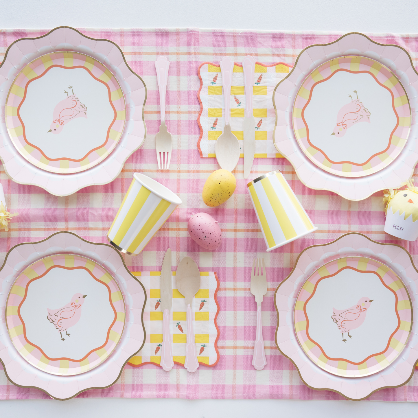 Table setting with pink and yellow plates, cups, and cutlery on a pink checkered tablecloth.