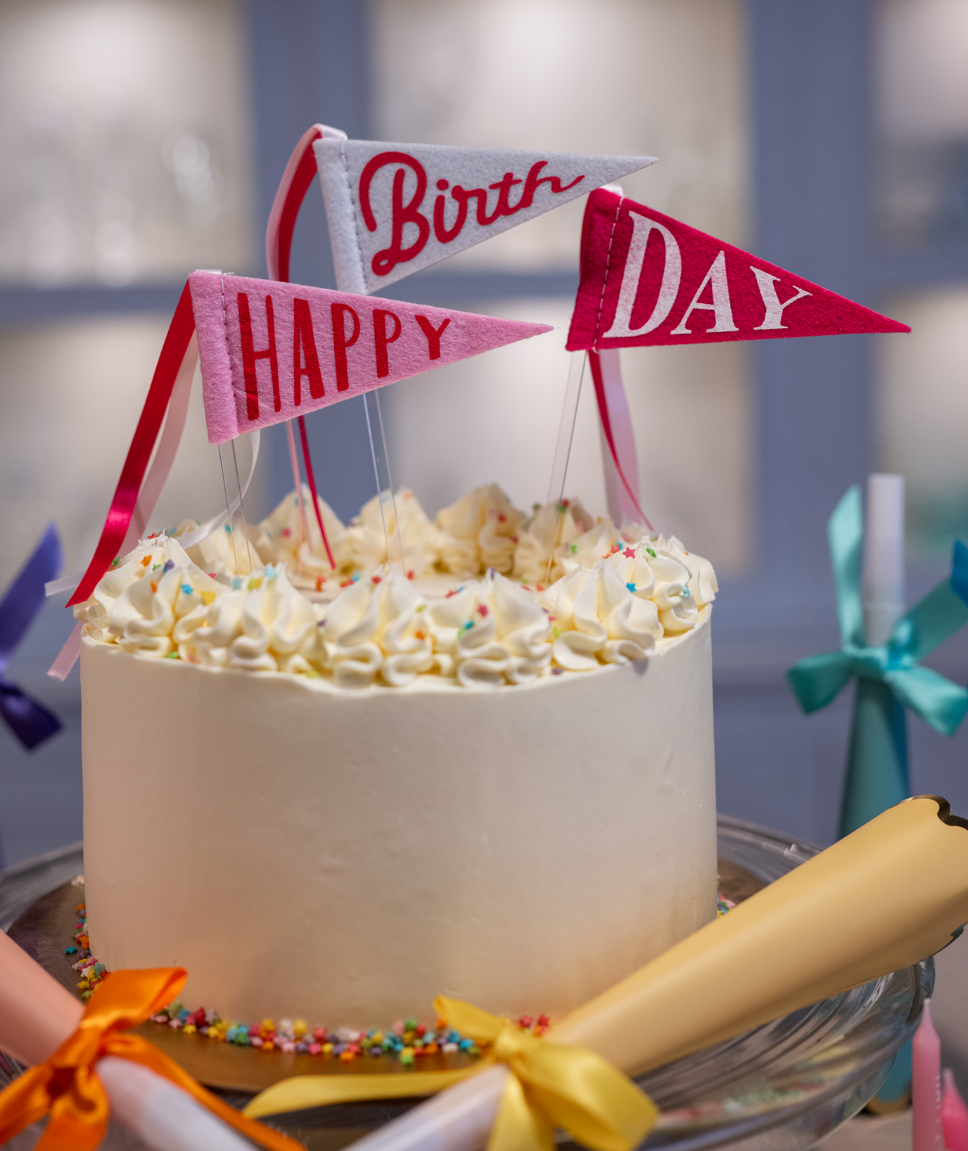 A round birthday cake with white frosting and piped cream on top, decorated with colorful sprinkles and three small pennant flags reading 'Birth', 'HAPPY', and 'DAY'.