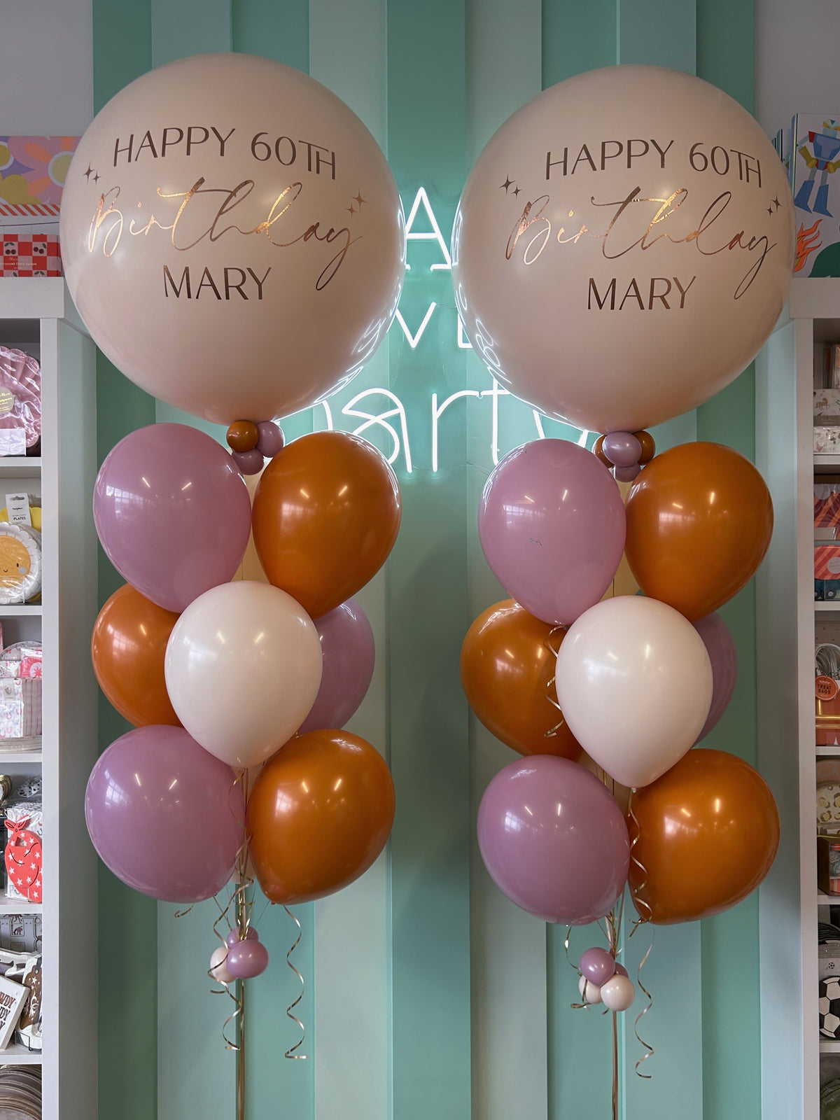 Close-up of two cream jumbo latex balloons personalized with Happy 60th Birthday Mary in gold and brown lettering, paired with mauve, orange and ivory helium balloon bouquets.