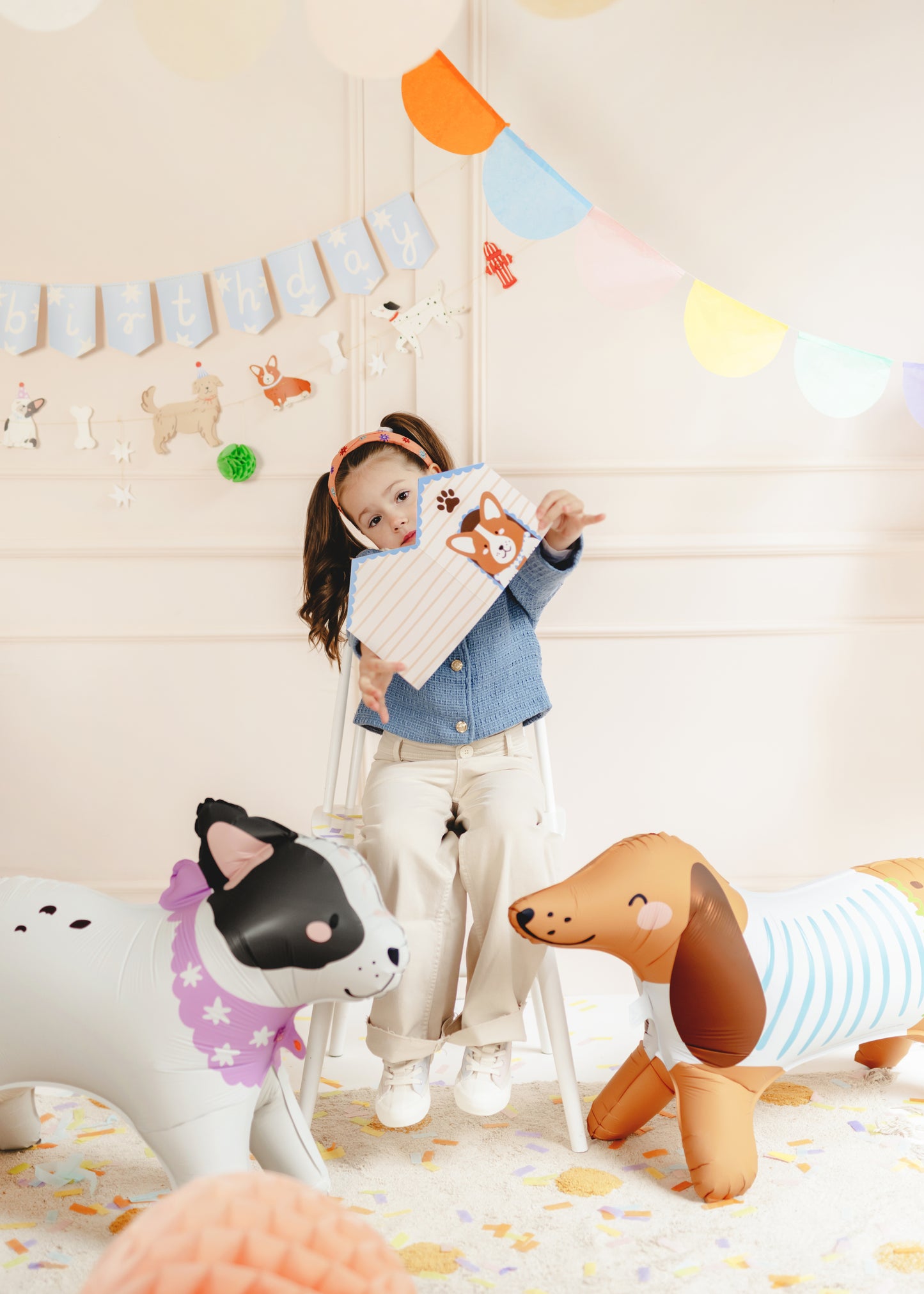 Child holding a dog-themed party favour box surrounded by inflatable puppy balloons, confetti, and a “happy birthday” banner.