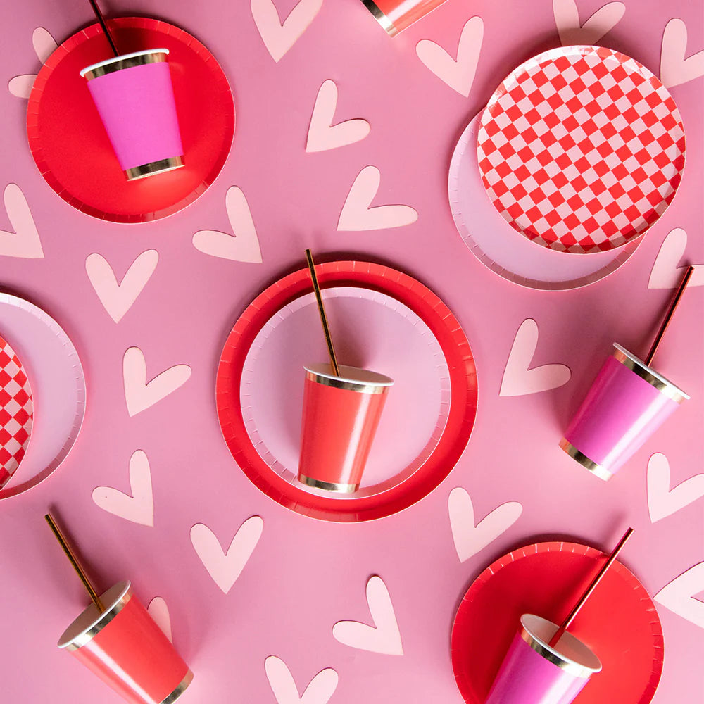 Flat lay of pink and red party plates, checkered designs, and metallic cups with straws on a heart-covered backdrop. Perfect for Valentine's Day or Galentineβs party decor.
