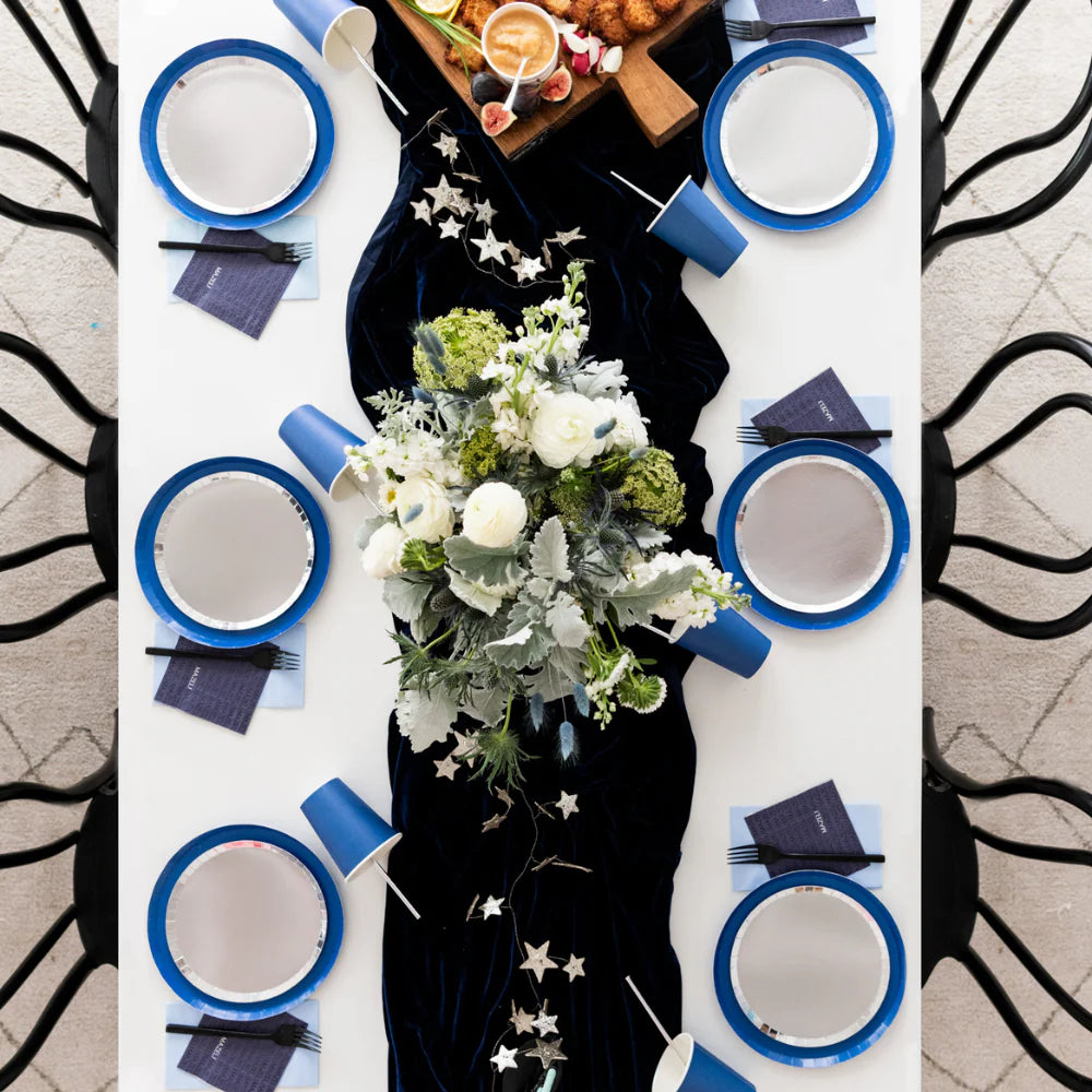 Elegant table setting featuring shiny silver dessert plates with royal blue borders, navy โMazelโ napkins, and a dark velvet runner accented with stars and white floral centerpieces.