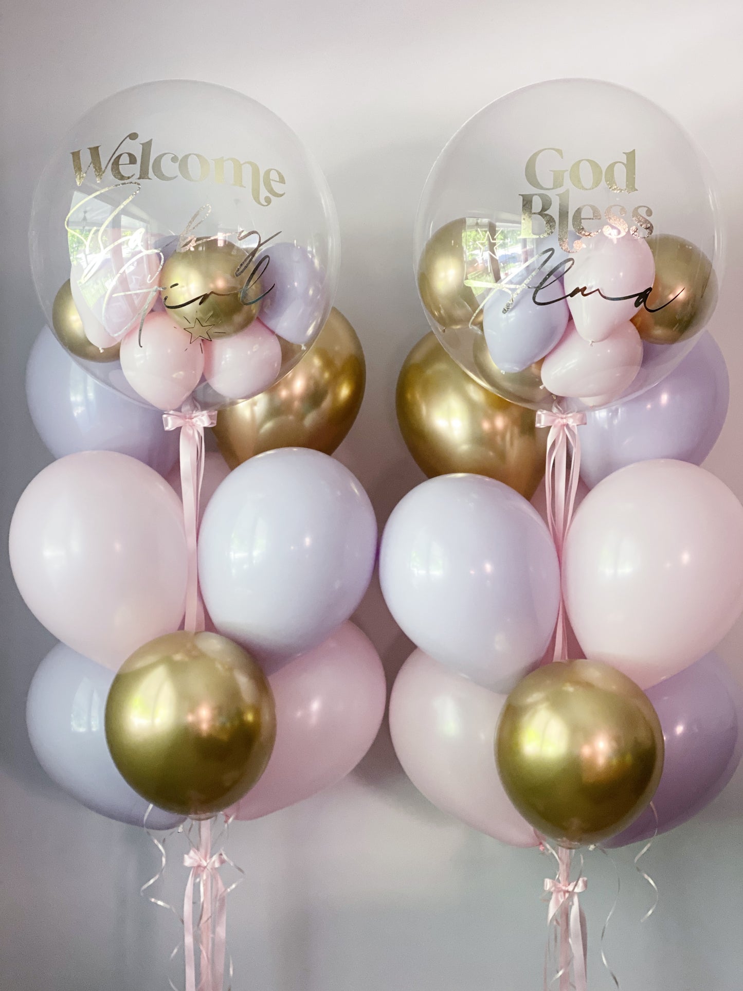 Two balloon arrangements with clear round balloons printed with 'Welcome Baby' and 'God Bless Baby,' filled with pastel pink, lavender, and metallic gold balloons, tied with pink ribbons against a light gray background.