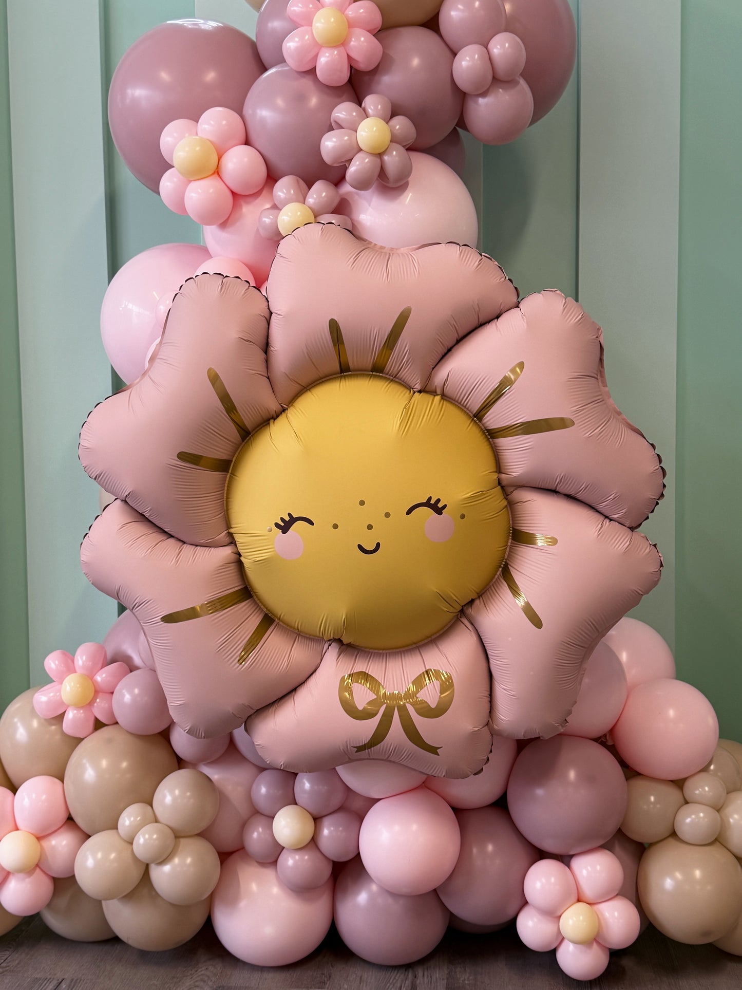 A decorative balloon arrangement featuring a large pink flower-shaped foil balloon with a smiling yellow sun face in the center, surrounded by smaller pink, beige, and yellow balloons against a pale green paneled backdrop.