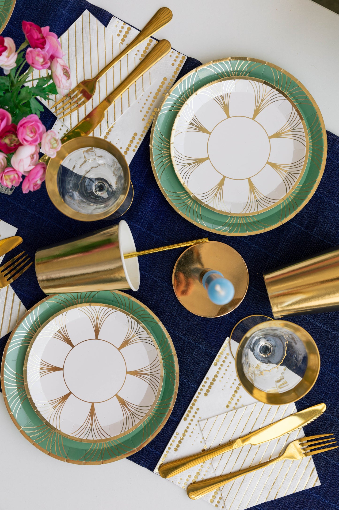 Elegant art deco-inspired table setting featuring white and gold dessert plates with green borders, gold flatware, gold cups, and striped napkins on a navy table runner with pink flowers.