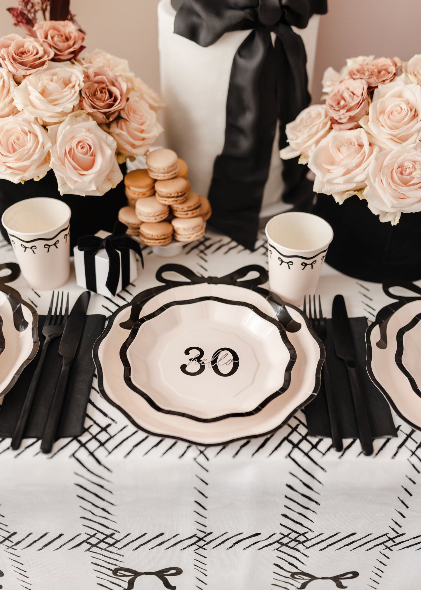Bow-themed 30th birthday table setting with cream and black paper plates, blush roses, and macarons.