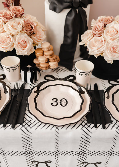 Bow-themed 30th birthday table setting with cream and black paper plates, blush roses, and macarons.