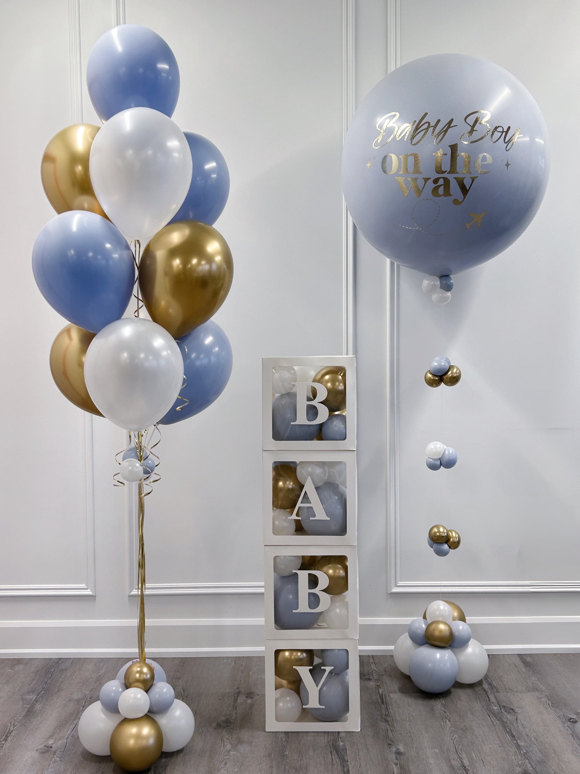 A baby shower decoration set featuring a cluster of light blue, white, and metallic gold balloons on the left, a large light blue balloon with gold foil text reading 'Baby Boy on the way' on the right, and a stack of three clear blocks displaying the letters 'B', 'A', 'B', 'Y' with gold and blue balloons inside. 