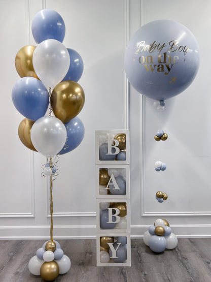 A baby shower decoration set featuring a cluster of light blue, white, and metallic gold balloons on the left, a large light blue balloon with gold foil text reading 'Baby Boy on the way' on the right, and a stack of three clear blocks displaying the letters 'B', 'A', 'B', 'Y' with gold and blue balloons inside. 