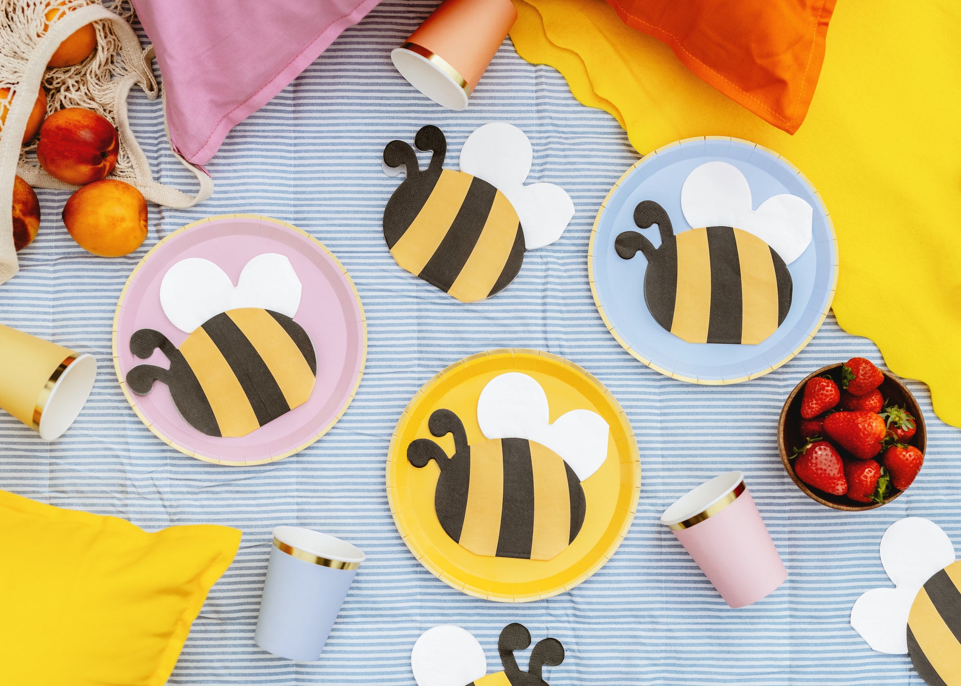 Bee-themed napkins and cups on a striped tablecloth with strawberries.