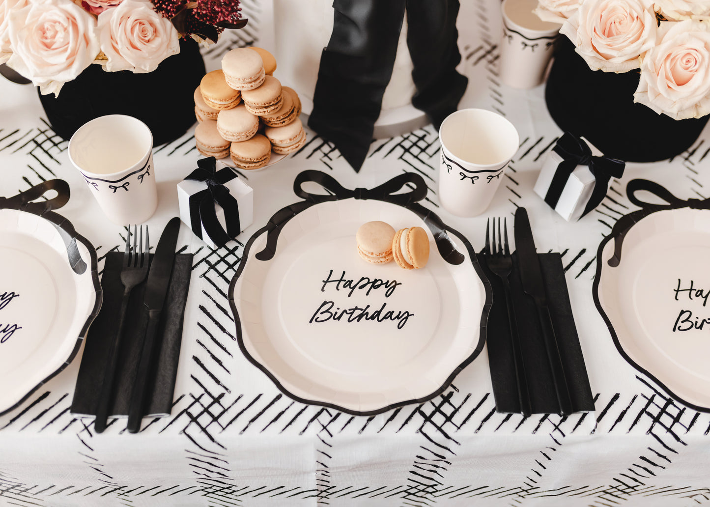Chic black and cream bow-themed birthday table setup with Happy Birthday plates, macarons, and rose centerpieces. Toronto party shop, ships Canada-wide.