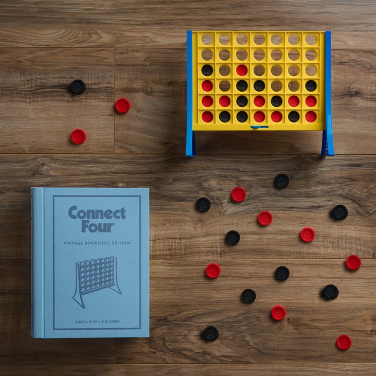 Connect Four game set on a wooden surface with book case. Sold in Toronto at Confetti My Party and ships Canada wide.