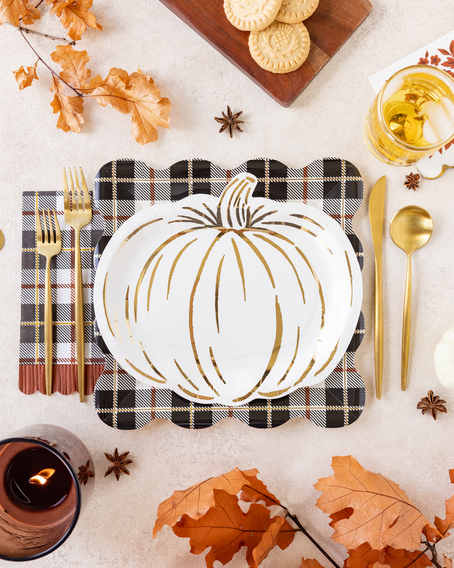 A styled autumn table setting featuring a white pumpkin-shaped plate with gold leaf accents, placed on a black and white plaid napkin with gold flatware. The scene is decorated with orange fall leaves, star anise, a candle, cookies on a wooden board, and a glass of iced tea on a light beige surface.