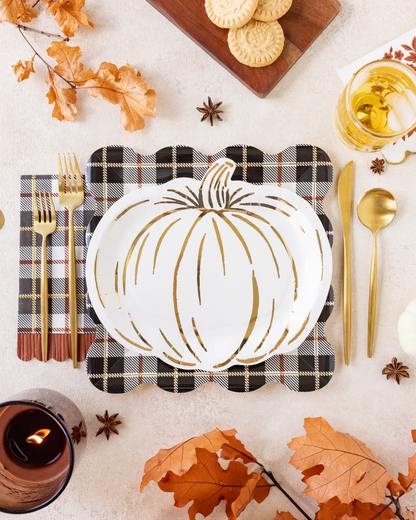 A styled autumn table setting featuring a white pumpkin-shaped plate with gold leaf accents, placed on a black and white plaid napkin with gold flatware. The scene is decorated with orange fall leaves, star anise, a candle, cookies on a wooden board, and a glass of iced tea on a light beige surface.