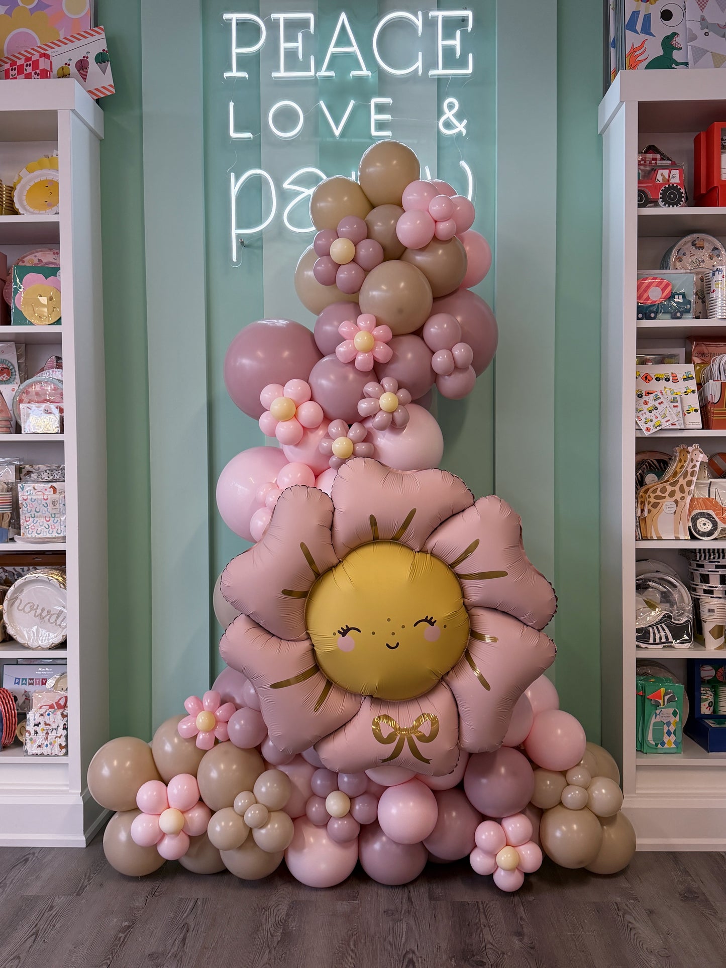 A decorative balloon garland arrangement shaped like a smiling sunflower, with pastel pink, dusty rose, beige, and yellow balloons, displayed in a nursery or playroom setting with shelves and a neon sign in the background.