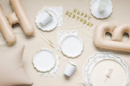 Flat lay of an elegant gold and white birthday party setup with scalloped plates, white cups with gold rims, "Happy Birthday" candles, and large nude letter balloons