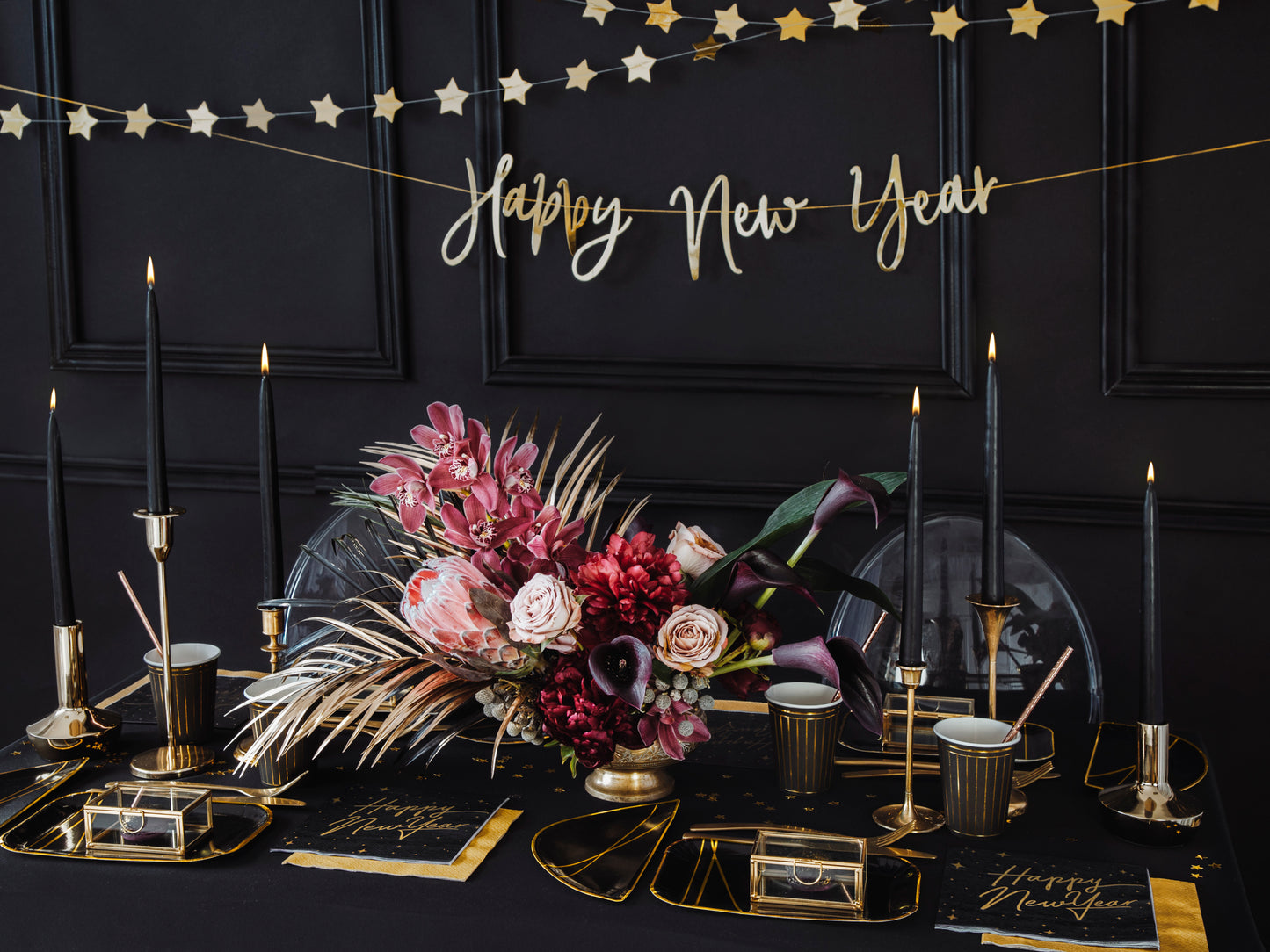 Elegant New Year’s Eve table with black taper candles in gold holders, floral centrepiece, and black and gold party decor.