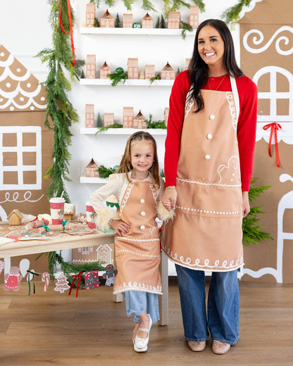 Woman and child wearing matching aprons in a festive setting with gingerbread house decorations.