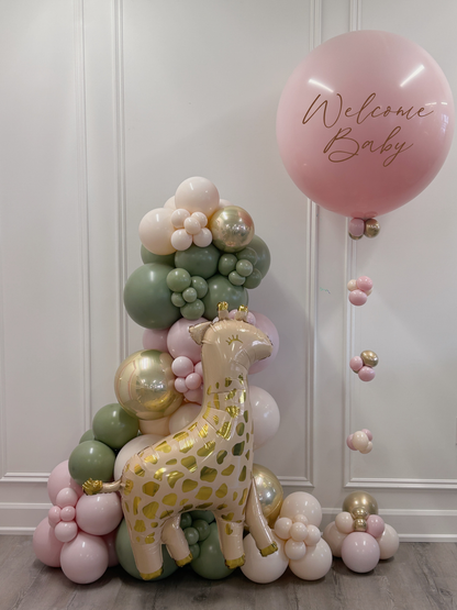 A baby shower decoration setup featuring a large inflatable giraffe balloon with gold foil spots, surrounded by a pastel balloon garland in pink, sage green, cream, and metallic gold. A large pink balloon with 'Welcome Baby' in brown script floats to the right against a white paneled wall.