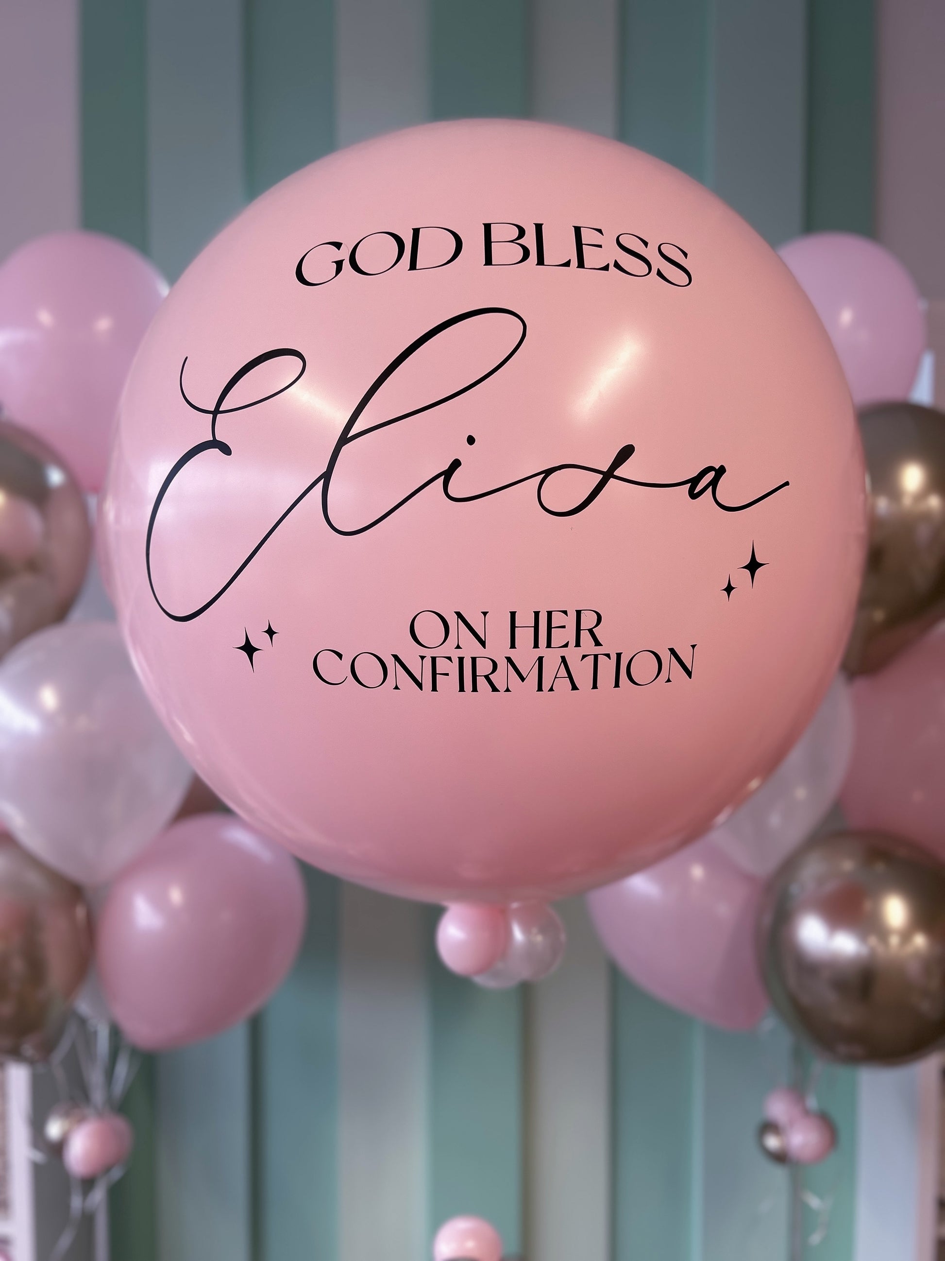 Pink balloon with 'GOD BLESS Elisa ON HER CONFIRMATION' text among other balloons against a striped background