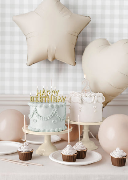 Neutral-toned birthday setup featuring two vintage-style cakes with piped frosting and gold "Happy Birthday" and number 2 candles, surrounded by nude star and heart balloons, cupcakes, and beige tableware