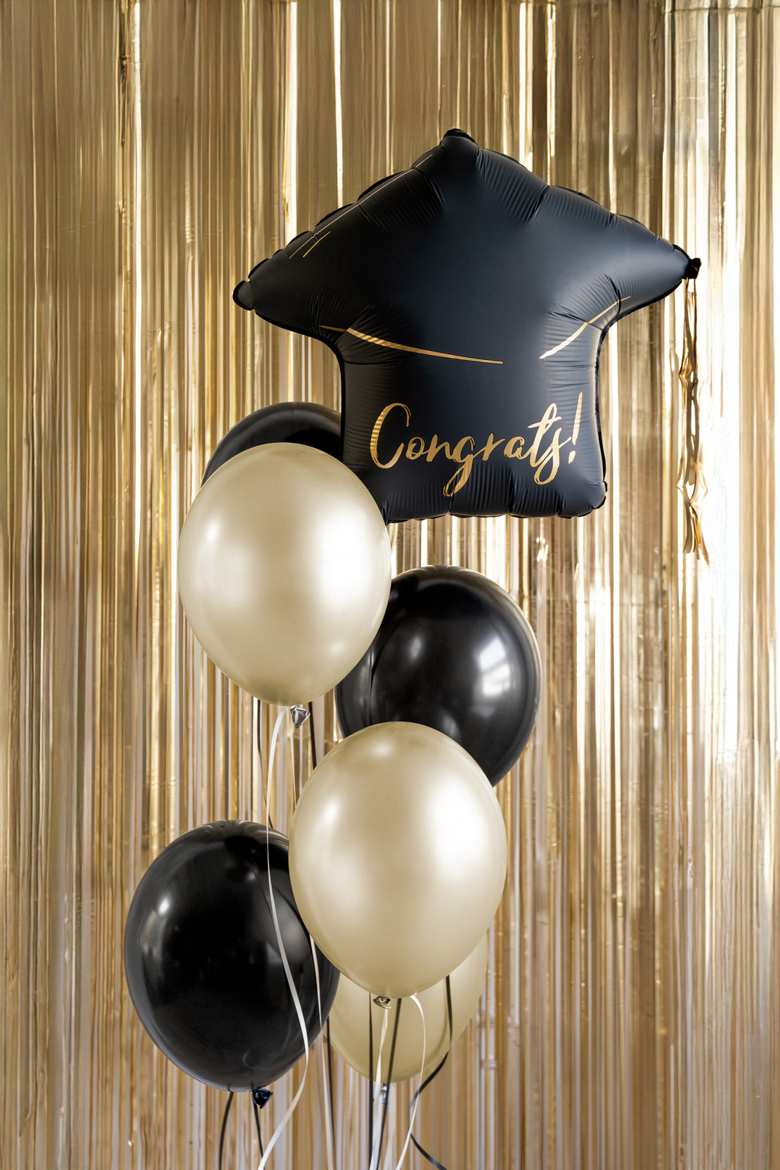 Black and gold graduation balloon bouquet with a “Congrats!” cap balloon, styled in front of a shiny gold foil fringe curtain backdrop. Perfect for grad party decorations or photo booth setup.