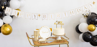 Gold Happy Birthday cake topper displayed on a birthday cake with black, white, and gold party decorations, available at our Toronto party shop.
