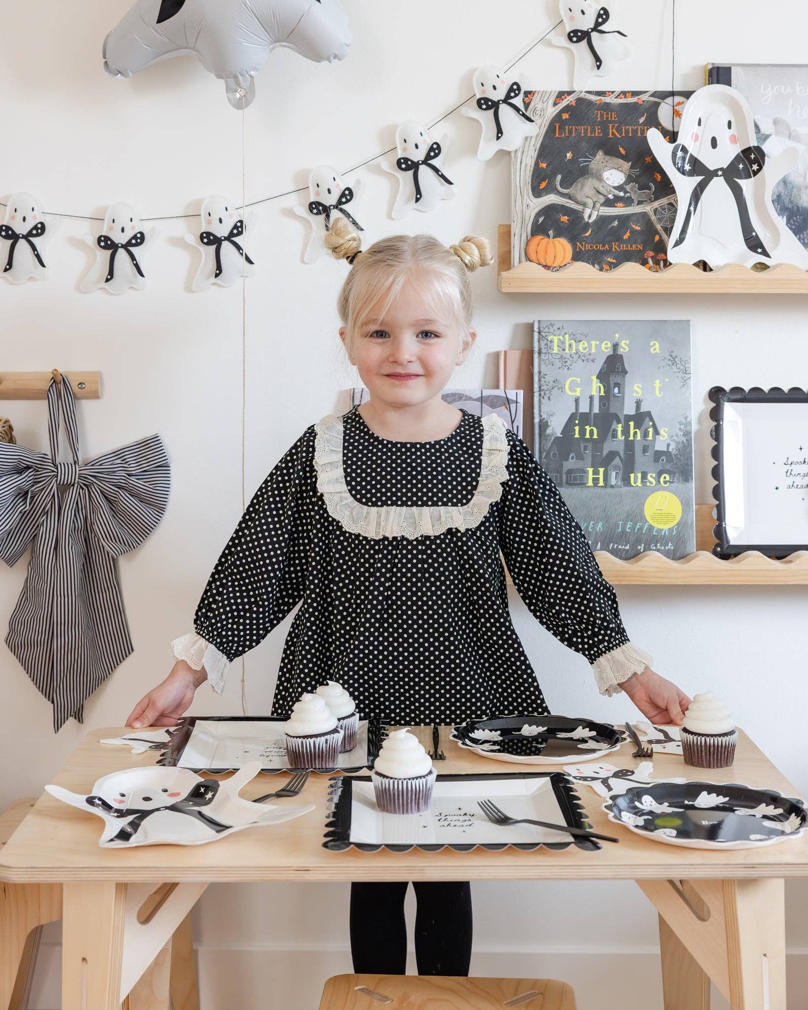 Child in a black polka dot dress standing at a table with Halloween-themed decorations.