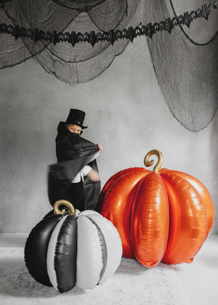 Person in a black outfit with a top hat sitting on a large inflatable pumpkin against a gray wall with spider web decorations.