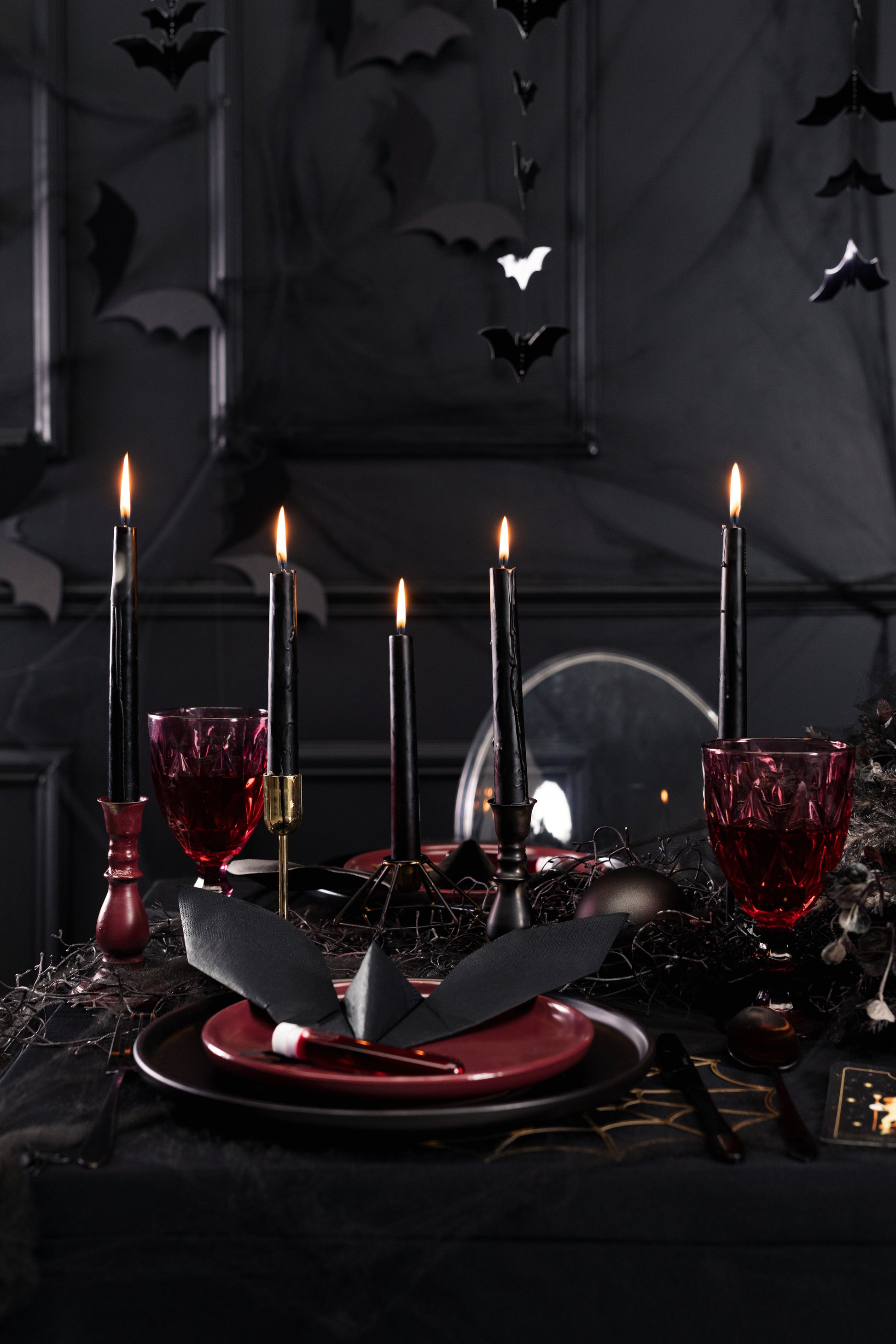 Halloween table setting with lit black taper candles, gothic tableware, red glasses, and bat decorations.