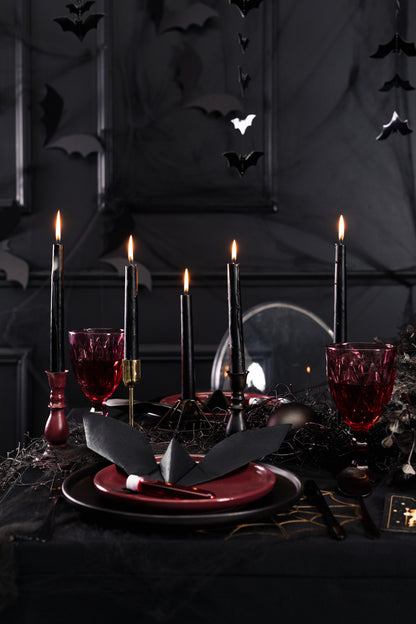 Halloween table setting with lit black taper candles, gothic tableware, red glasses, and bat decorations.