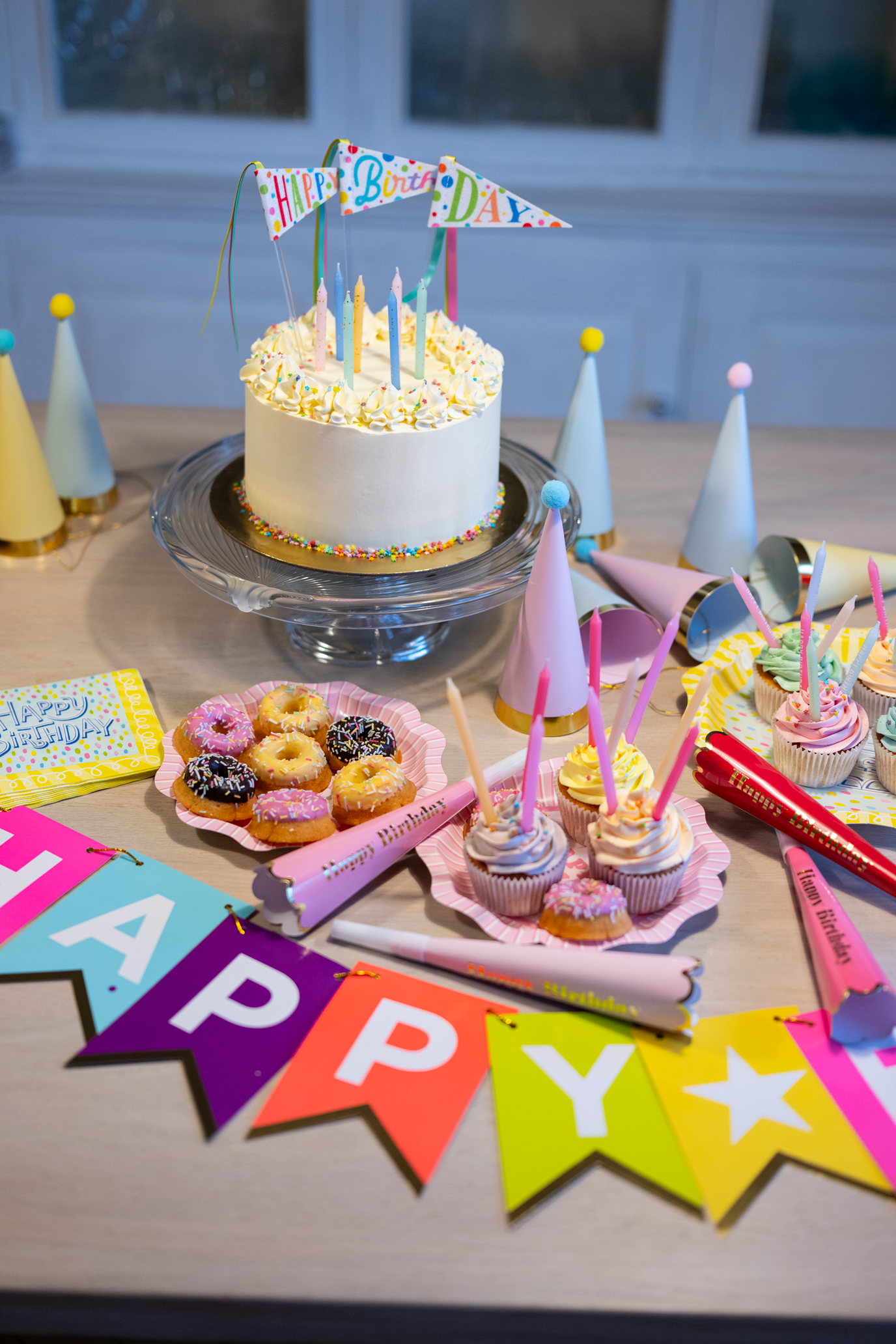 Birthday cake with lit candles, colorful decorations, and a 'Happy Birthday' banner on a table.