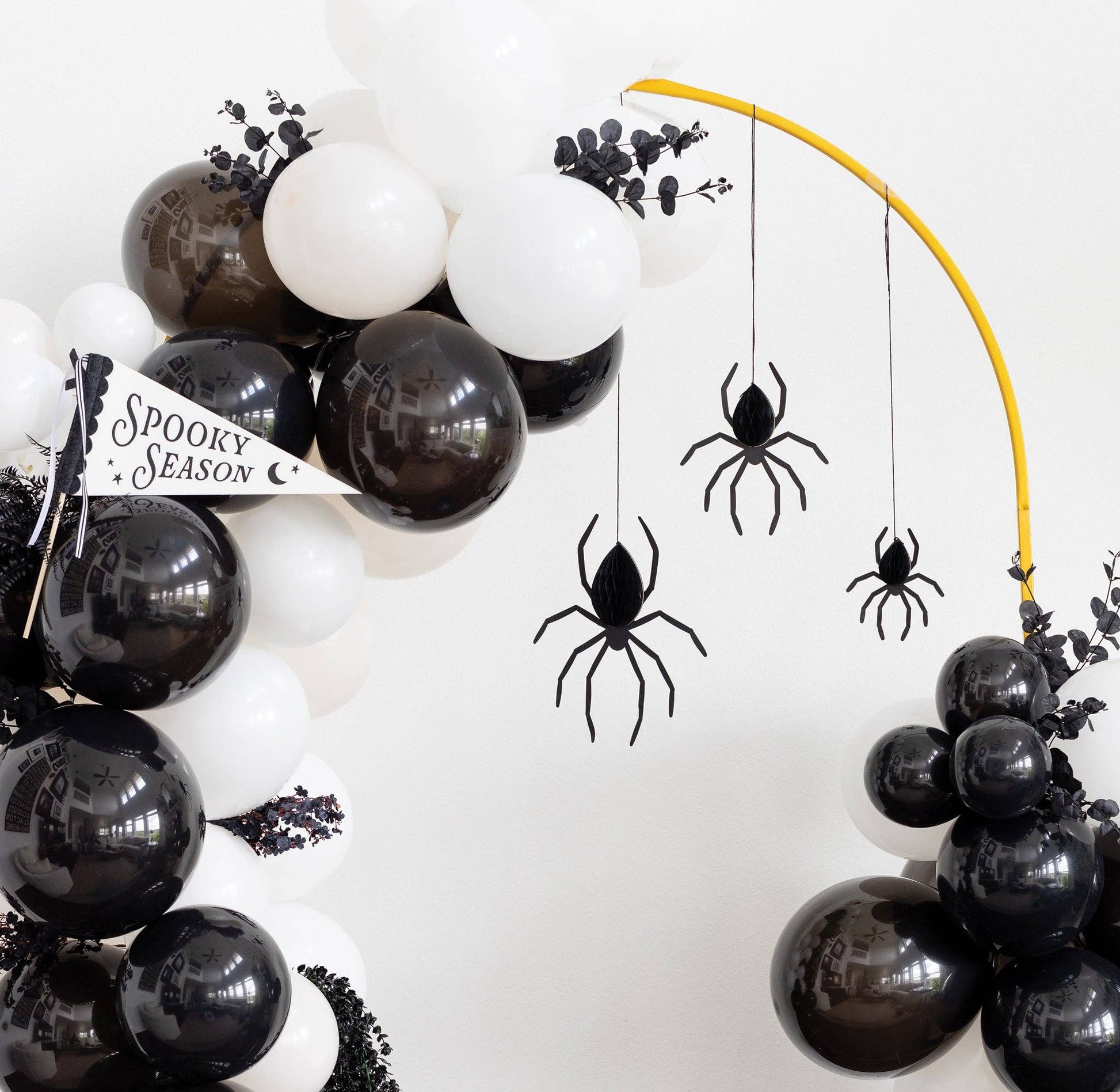 Halloween balloon garland decoration with black and white balloons, black spider cutouts hanging from a yellow arch, and a small 'Spooky Season' banner.