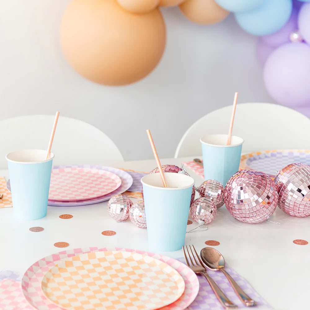 Pastel party table featuring peach and pink checkered plates, blue cups with straws, pink disco balls, and balloon garland.