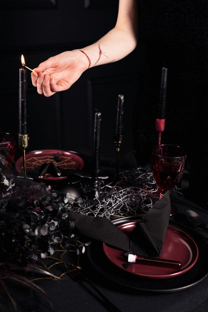 Hand lighting a matte black taper candle on a moody Halloween table with dark tableware and red details.
