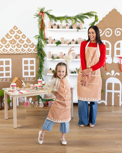 Two people wearing matching aprons in a festive kitchen setting with gingerbread house decorations.