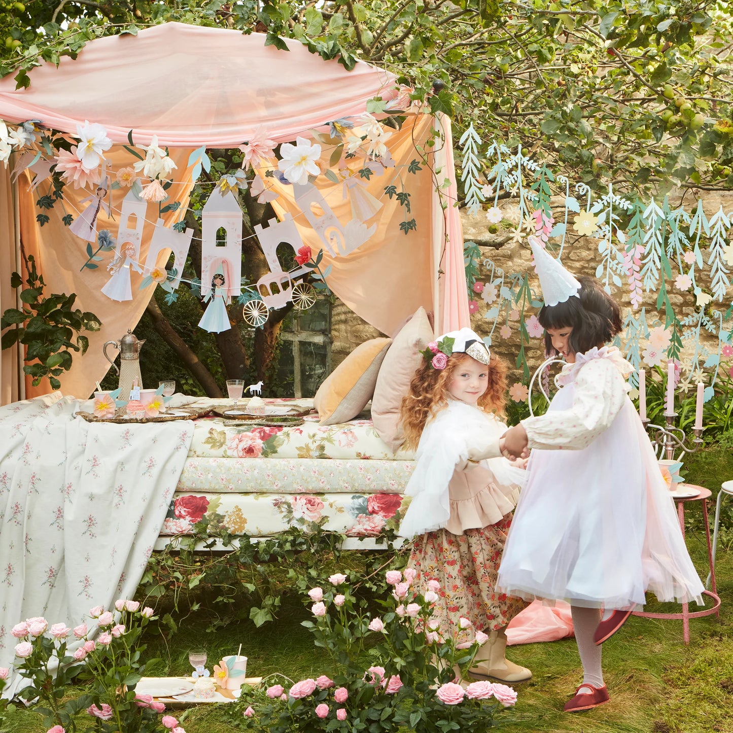 Two children playing at a fairy tale-themed outdoor party featuring Meri Meri castle and princess garland hanging on a peach canopy.