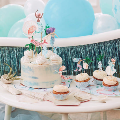 Table with a cake and cupcakes decorated with mermaid-themed toppers, surrounded by balloons and a water-themed backdrop.