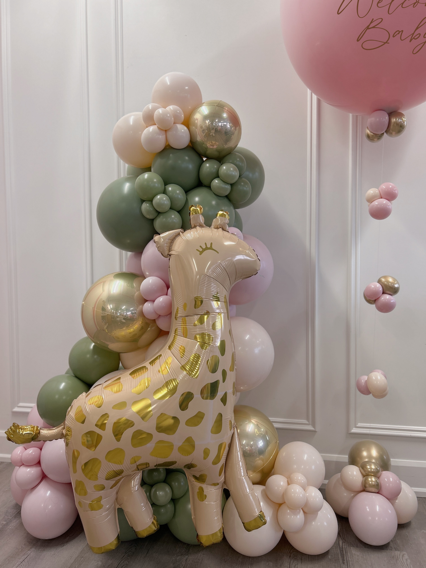 A baby shower decoration setup featuring a large metallic gold and beige foil giraffe balloon in front of a green, pink, and gold balloon garland, with a pink 'Welcome Baby' balloon in the corner.