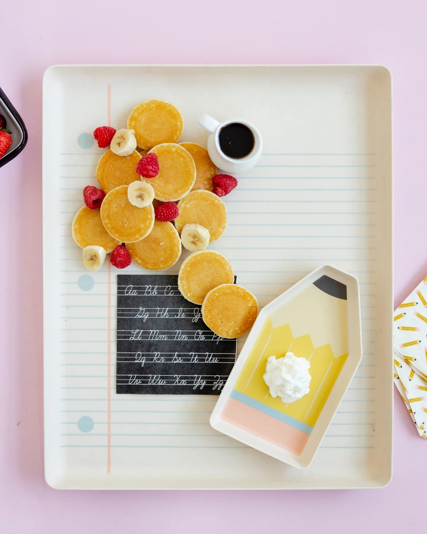 Platter of pancakes with fruit and a small pitcher of syrup on a pink background