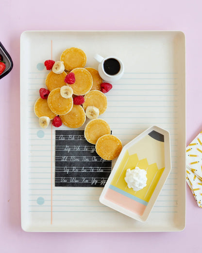 Platter of pancakes with fruit and a small pitcher of syrup on a pink background