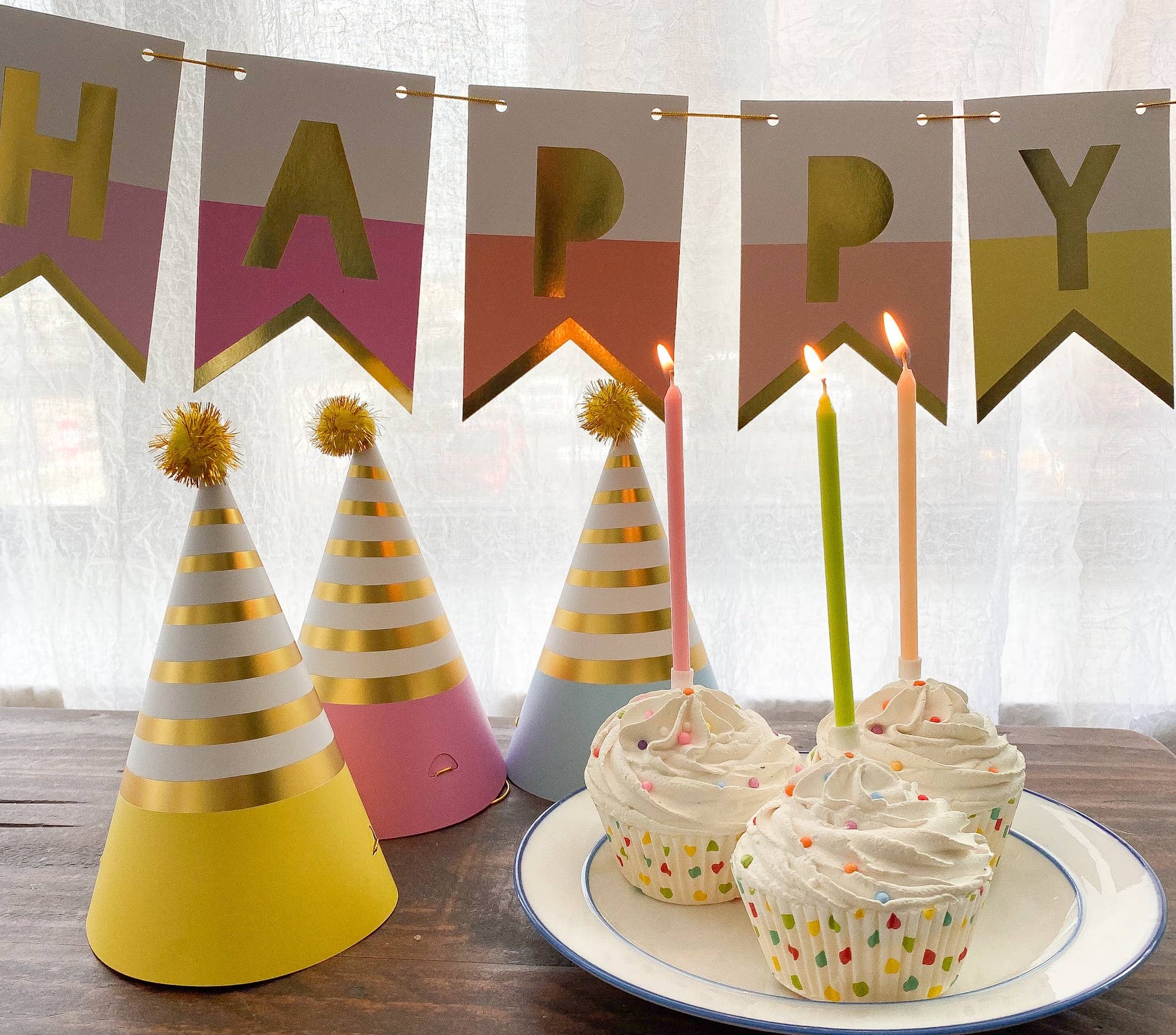 Birthday-themed items including colorful party hats and cupcakes with lit candles on a wooden table.
