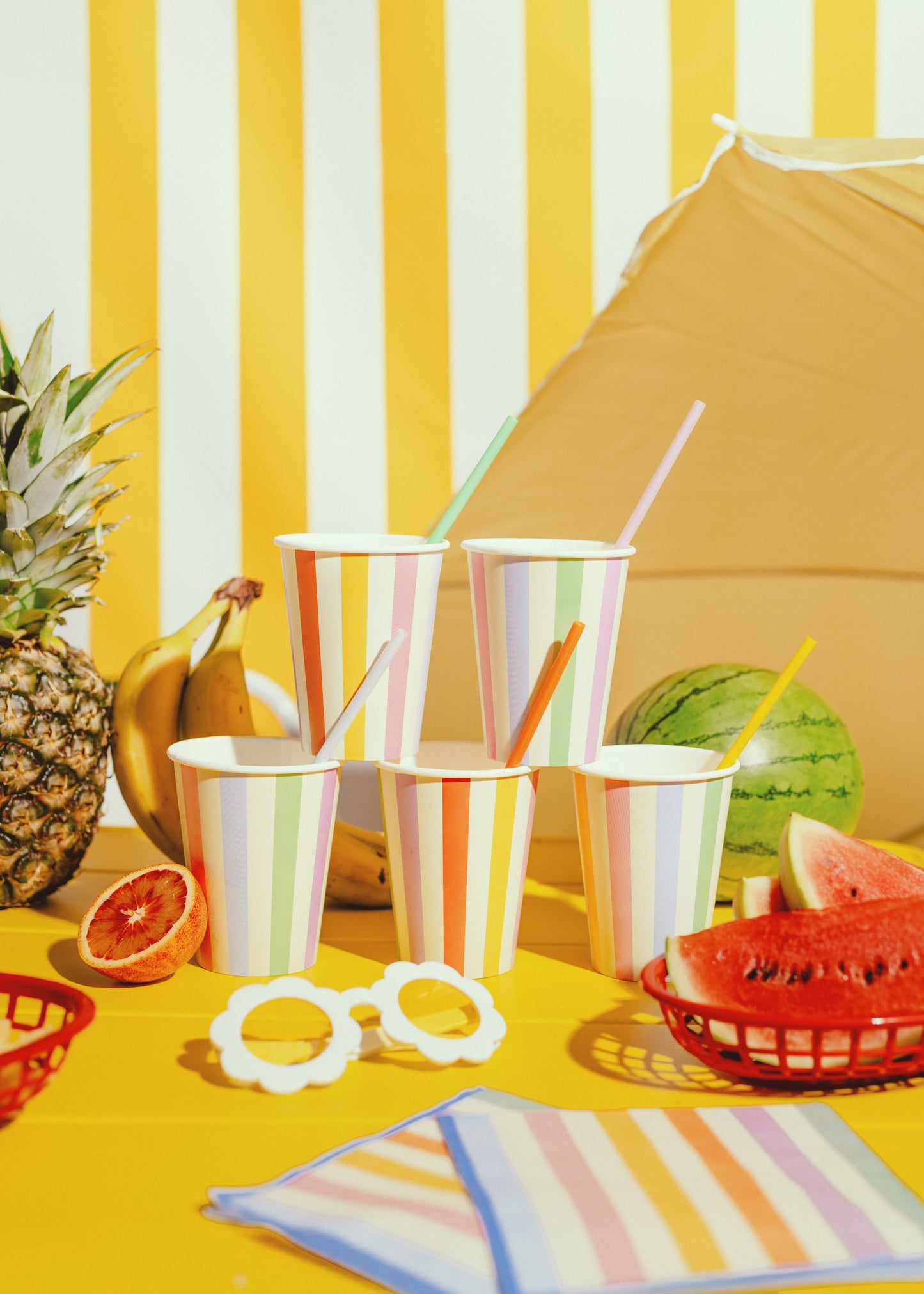 Colorful outdoor setting with striped tablecloth, cups, straws, and fruits on a yellow surface.