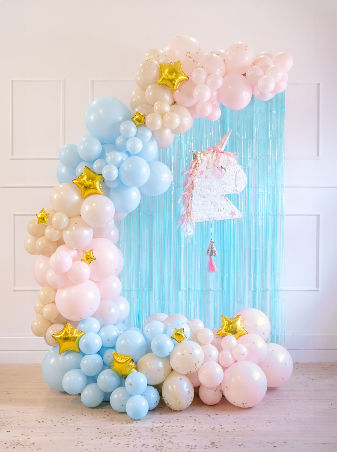 Pastel balloon garland in blue, blush, and cream with gold star balloons, styled around a sky blue foil fringe curtain and unicorn piñata. Perfect for a whimsical kids’ birthday or unicorn-themed party backdrop.