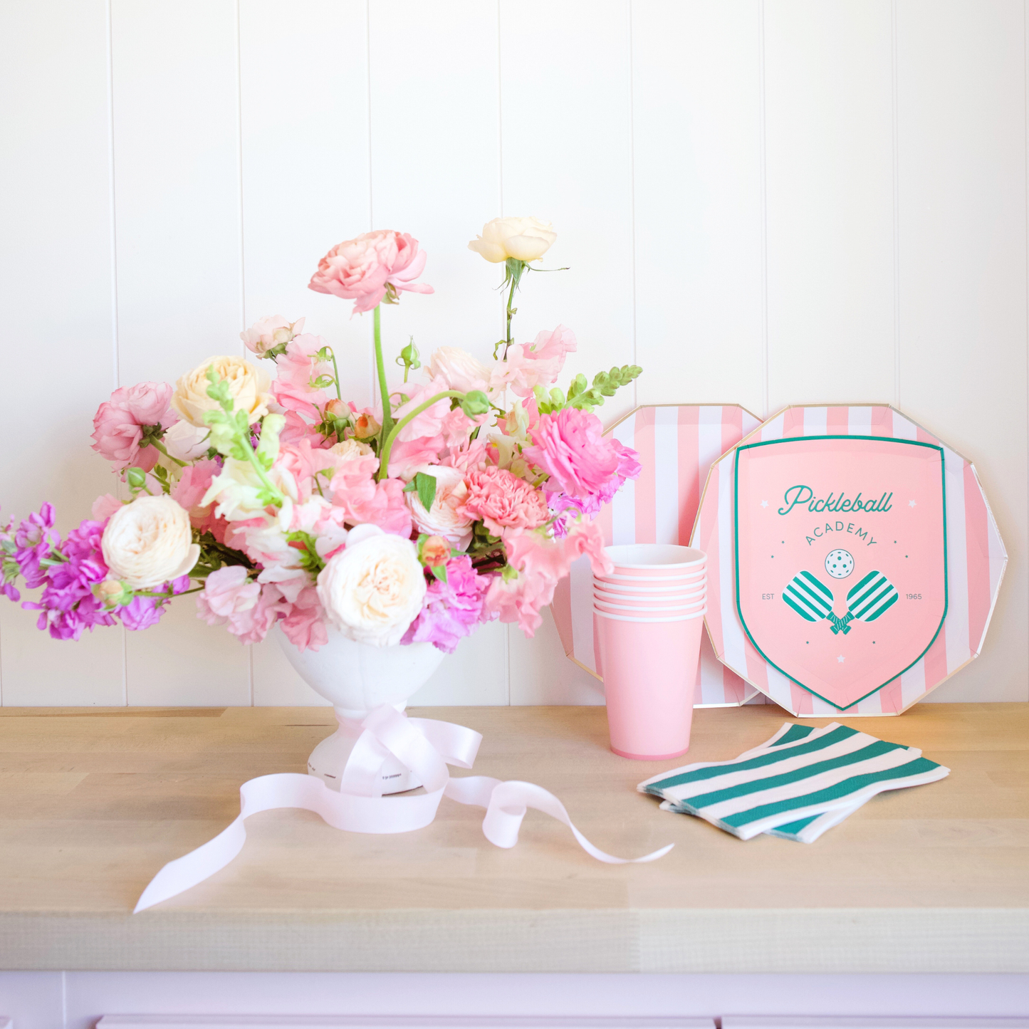 Floral arrangement with pink and white flowers, striped cups, and a pink and green party hat on a wooden surface.
