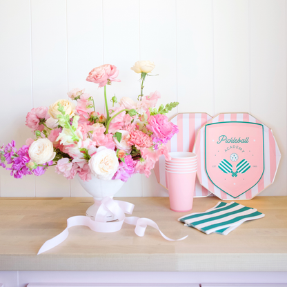 Floral arrangement with pink and white flowers, striped cups, and a pink and green party hat on a wooden surface.