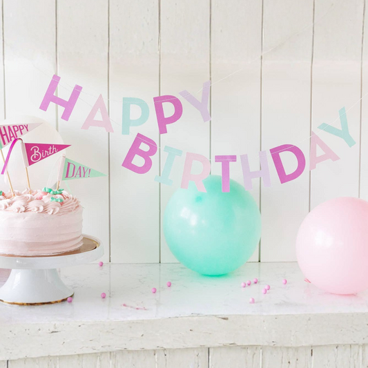Birthday cake with pink frosting, balloons, and a 'Happy Birthday' banner on a wooden surface.